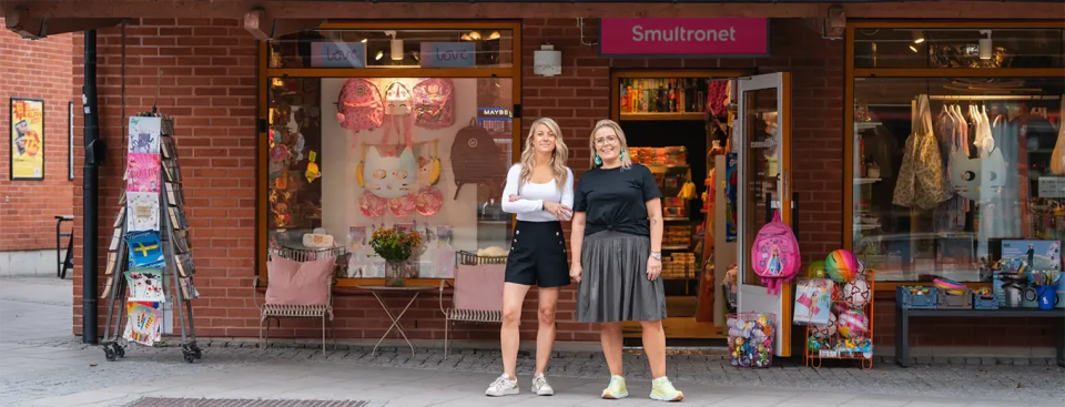 Två glada kvinnor i sommarkläder utanför en butikslokal.