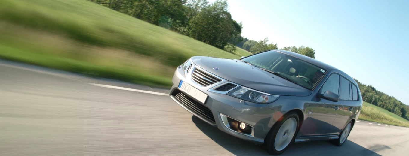 En grå Saab kör längs en landsväg omgiven av gröna fält och klarblå himmel, vilket ger en känsla av rörelse och hastighet