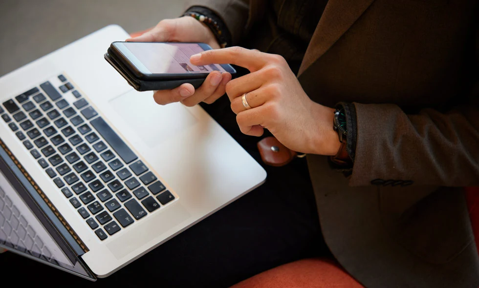 En person håller en smartphone i ena handen och använder den andra för att interagera med en laptop på ett mörkt bord.