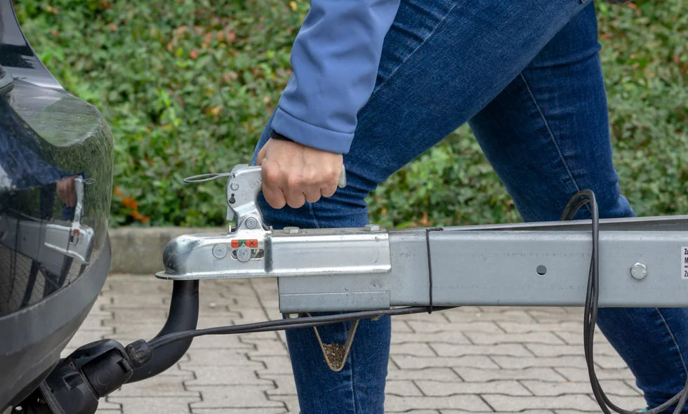 Person i blå jeans fäster en släpvagn till bilens dragkrok, omgiven av stenbelagd väg och grönska