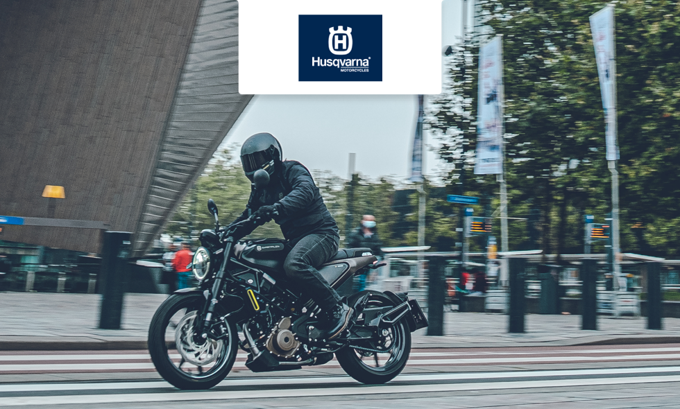 Förare på Husqvarna-motorcykel kör genom stadsmiljö i rörelse över ett övergångsställe.