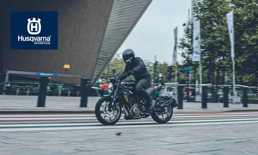 En förare i svart utrustning kör en klassiskt designad Husqvarna motorcykel på ett övergångsställe framför en modern byggnad
