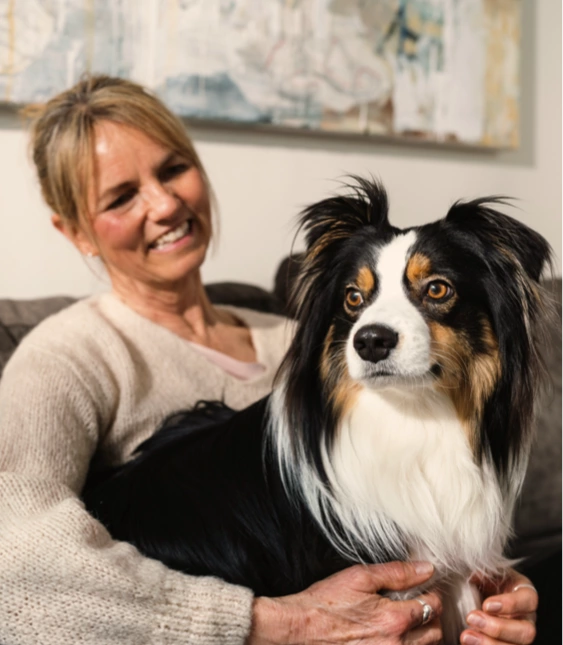 En glad kvinna sitter på en soffa och ler medan hon tittar på sin sittande svartvita hund med markanta färger och fluffig päls