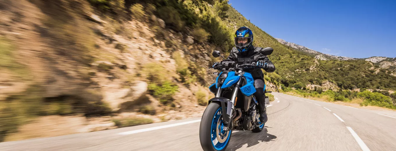 Motorcyklist kör på en solig landsväg med berg och naturlandskap i bakgrunden.