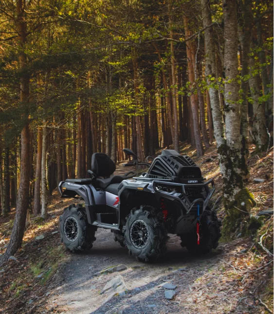 ATV parkerad på smal skogsstig i kuperad terräng, omgiven av höga träd och höstfärger, redo för körning i naturen.