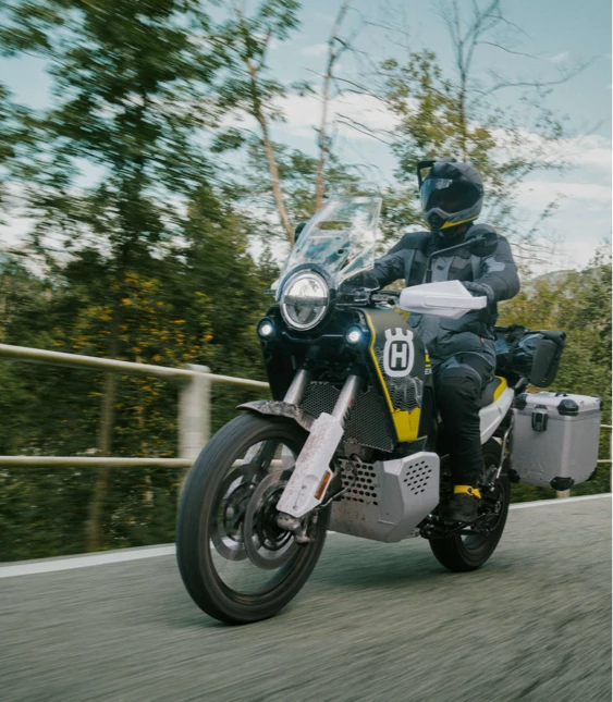 Motorcyklist i full skyddsutrustning kör på kurvig landsväg med berg och grönskande natur i bakgrunden.