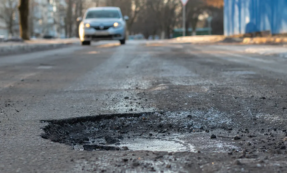 Närbild på ett djupt potthål fyllt med vatten i en asfalterad väg, med en oskarp bil som närmar sig i bakgrunden