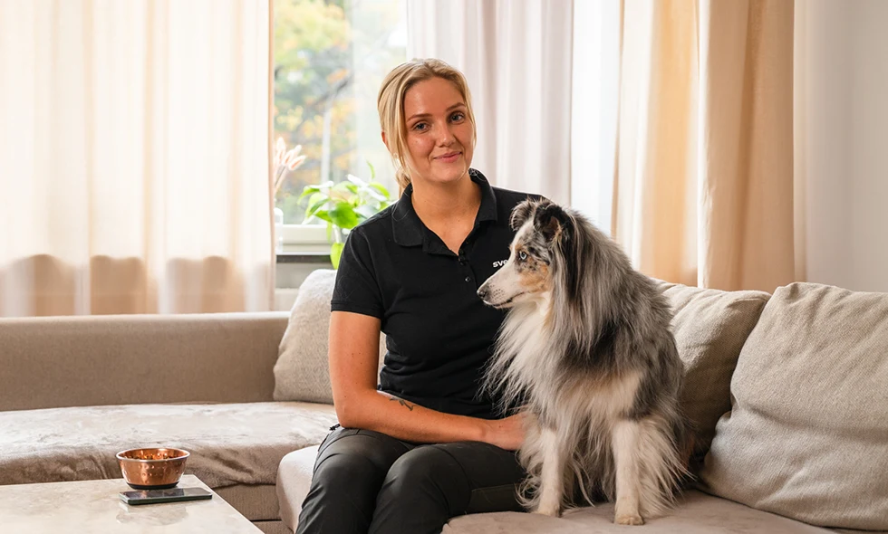 Kvinna i svart pikétröja sitter i en soffa med en långhårig collie bredvid sig i ett ljust vardagsrum.