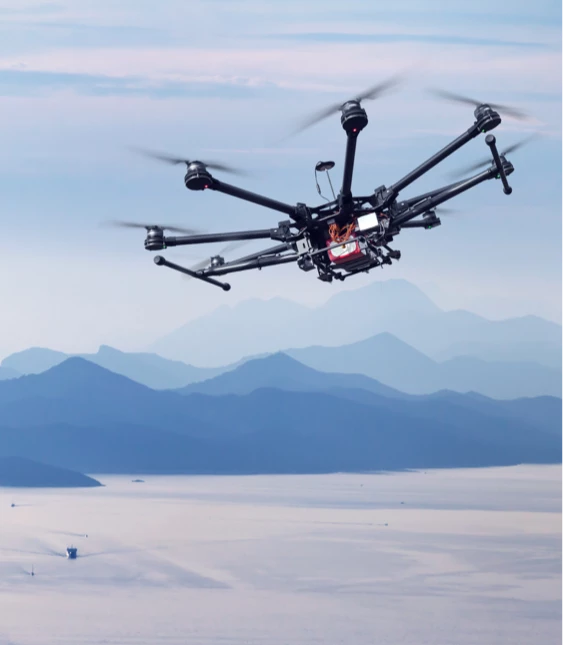 En stor drönare med svarta rotorblad flyger högt ovanför bergiga terränger i disig atmosfär, en teknologisk pryl i naturens lugn