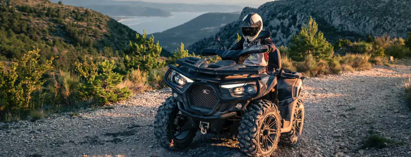 Person på svart ATV från Kymco stannar på grusväg i bergslandskap.