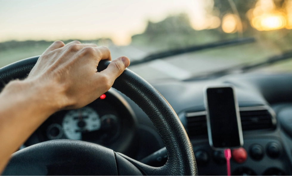 Hand på ratt i grynings- eller kvällsljus, suddig väg och mobil i hållare.