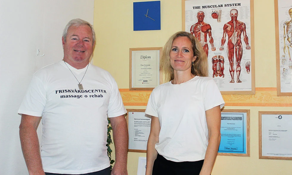 Två personer, en man och en kvinna, står framför en vägg med affischer och diplom, leende och klädda i vita T-shirts med texten 'FRISKVÅRDSCENTER massage & rehab'