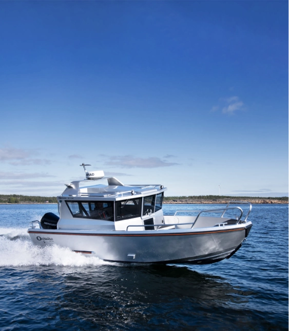 Aluminiumbåt av märket Ockelbo med kabin kör snabbt över en sjö med kustlinje och skog i bakgrunden.