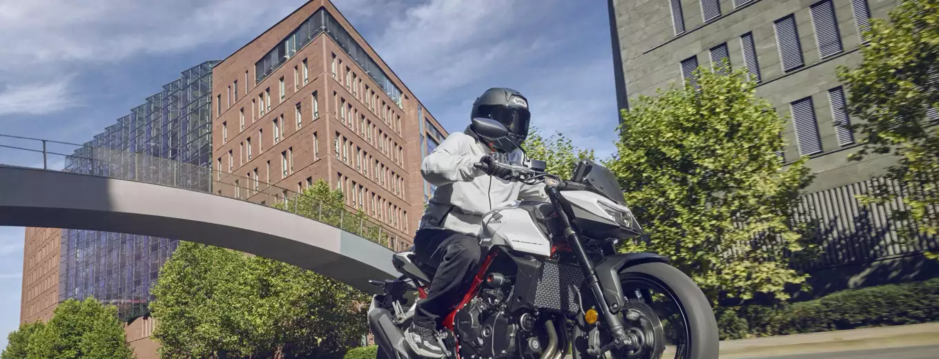 Motorcyklist kör på väg i stadsmiljö med grönskande träd i bakgrunden.