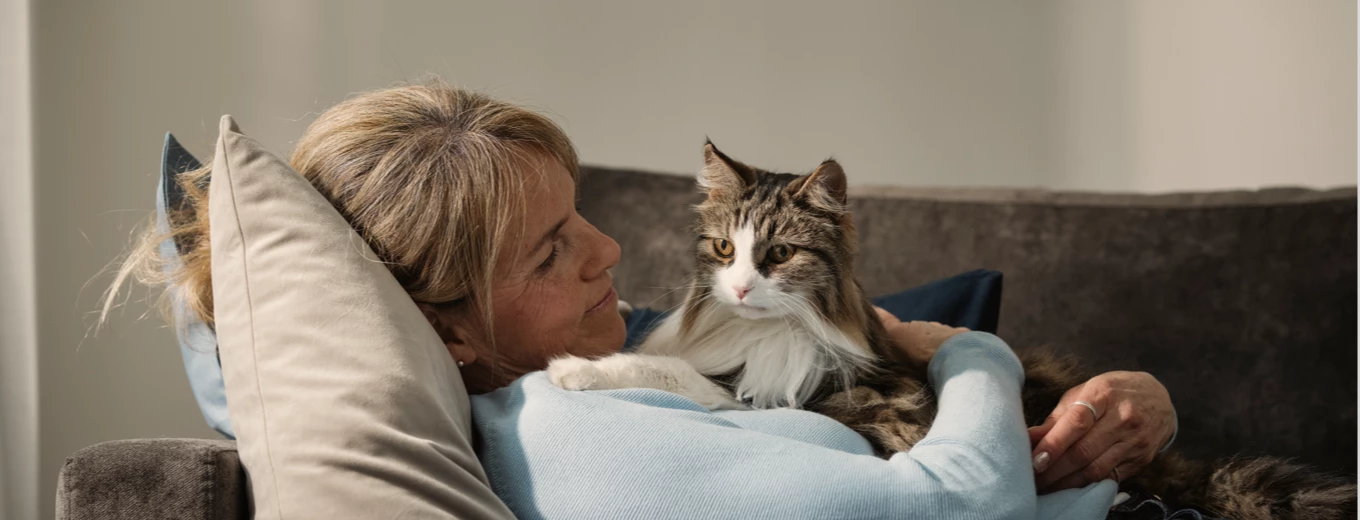 En kvinna ligger tillbakalutad på en soffa och håller om en fluffig katt, båda tittar mot kameran med avslappnade uttryck.