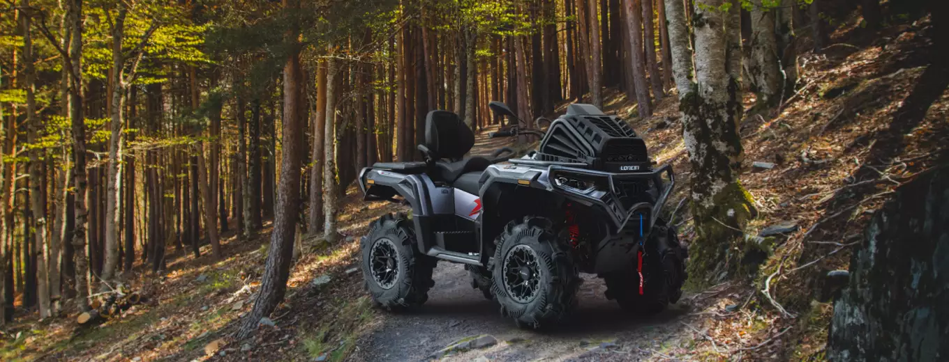ATV parkerad på smal skogsstig i kuperad terräng, omgiven av höga träd och höstfärger, redo för körning i naturen.