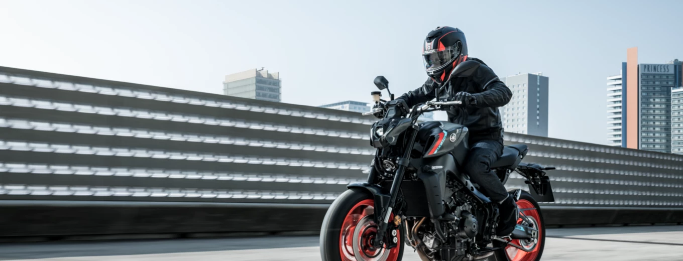En motorcyklist i full svart skyddsutrustning kör en svart motorcykel med röda detaljer på en upphöjd väg med stadssilhuetten i bakgrunden