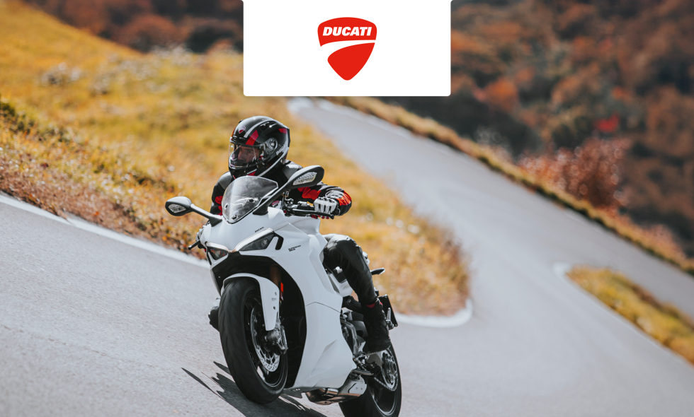 Förare av vit Ducati-motorcykel kör i en kurva längs slingrande landsväg i höstfärgat landskap.