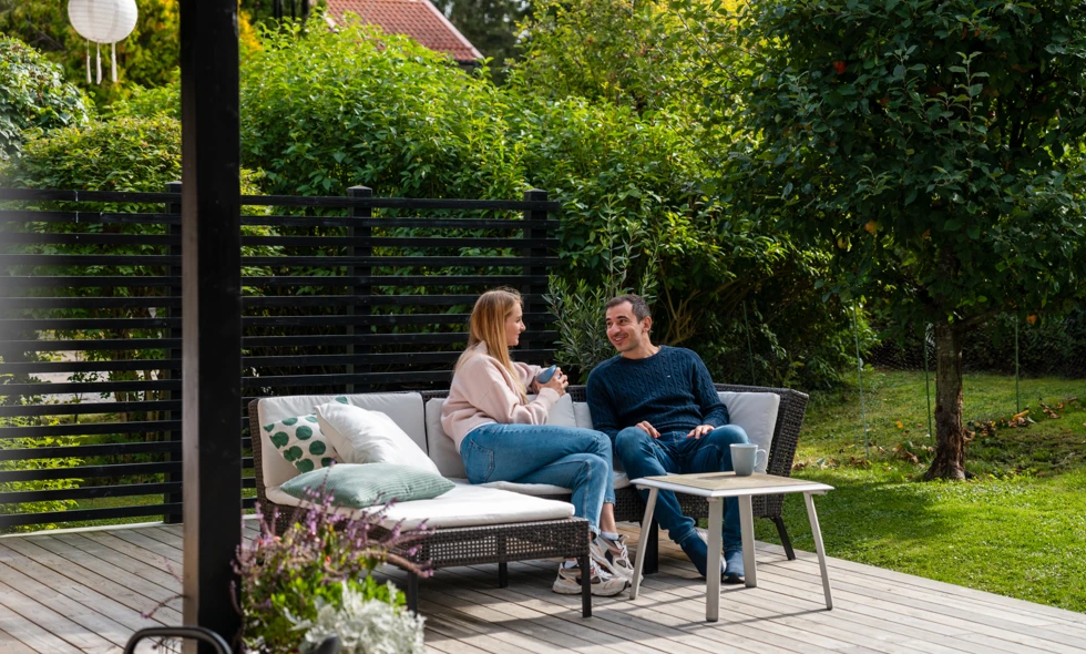 Två personer sitter i en soffgrupp och samtalar på en solig altan omgiven av grönska.