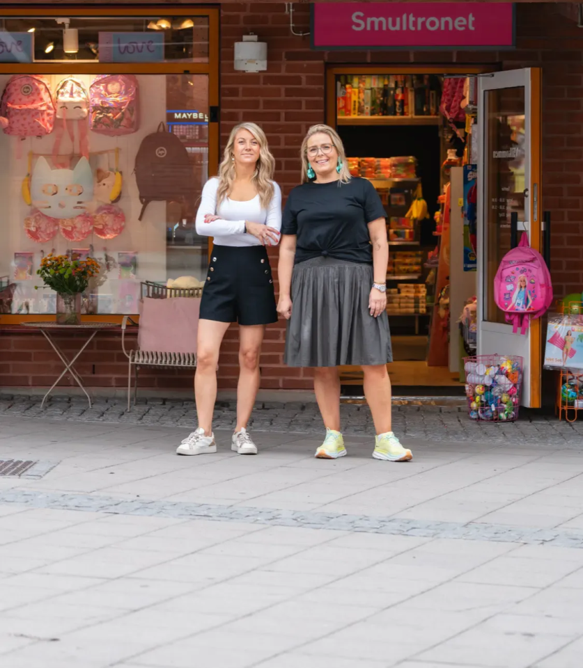 Två glada kvinnor i sommarkläder utanför en butikslokal.