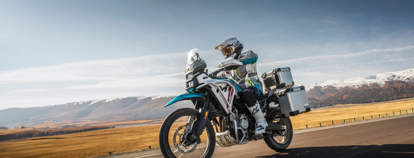 Motorcyklist med packväskor kör på landsväg med fjäll och öppet landskap i bakgrunden.