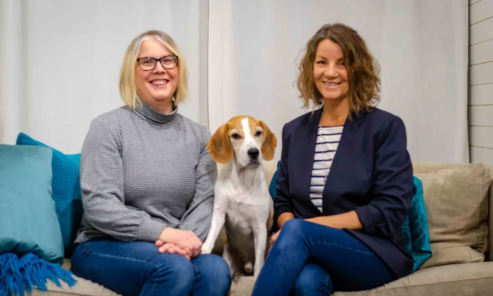 Två leende kvinnor sitter på en soffa med en beagle mellan sig)