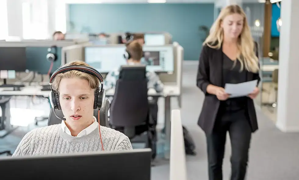  En ung man med headset vid dator i kontorslandskap, kvinna i bakgrunden