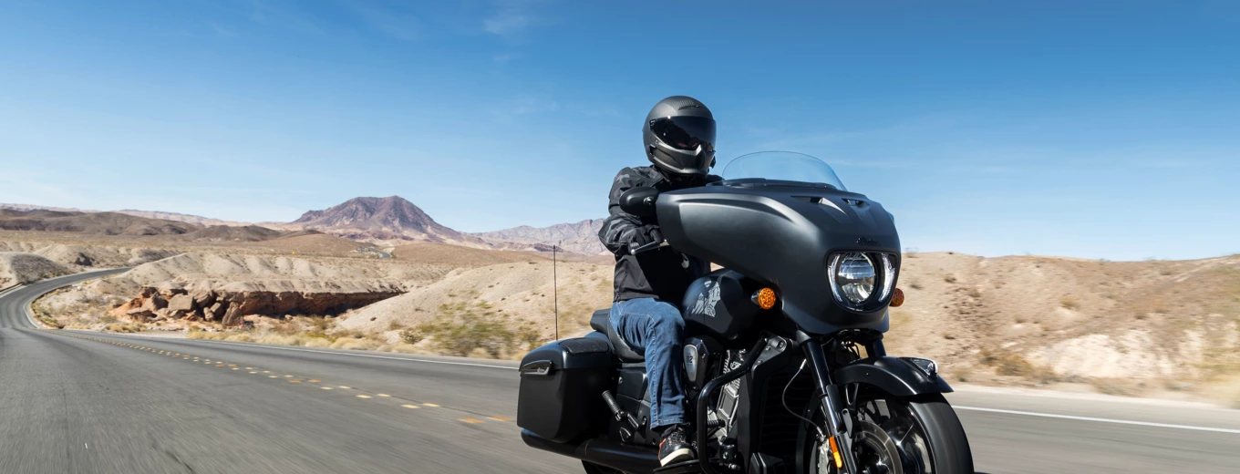 Motorcyklist kör på öde landsväg genom ökenlandskap med berg i bakgrunden under klarblå himmel.