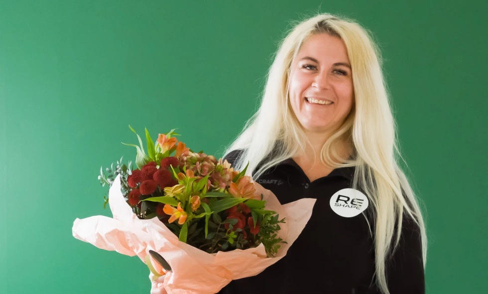 En kvinna i arbetskläder leende håller en bukett blommor framför en grön bakgrund)