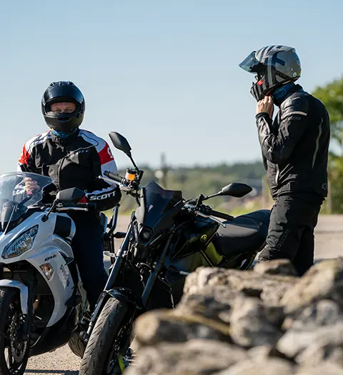 Två motorcyklister stannar på en utsiktsplats, en sittande på en svart motorcykel och den andra stående bredvid med hjälmen i handen