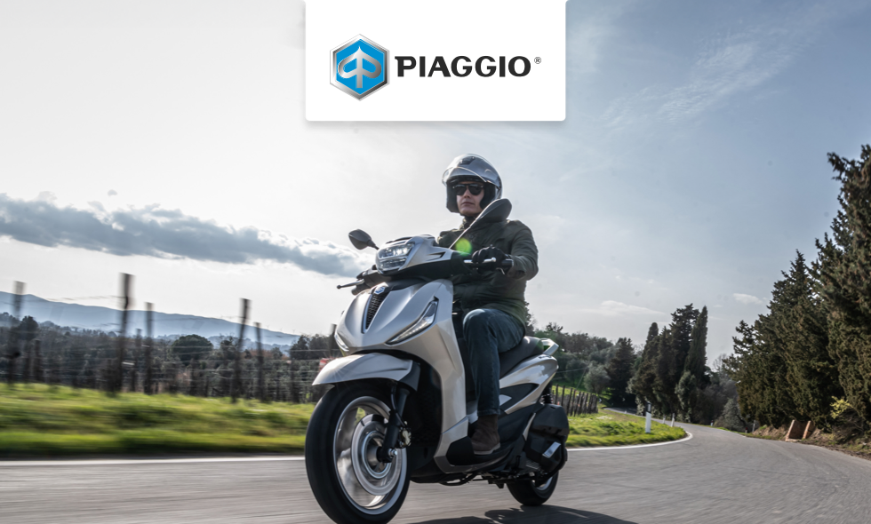 Person kör Piaggio-scooter på lantlig väg med träd och berg i bakgrunden.