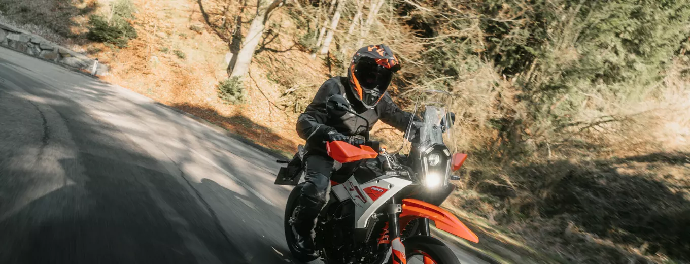 Motorcyklist i svart hjälm kör orange äventyrsmotorcykel genom kurva på skogsväg.