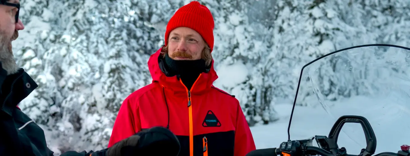 En man med skägg, orange mössa och röd jacka ler och tittar in i kameran, med en vintrig skogsbakgrund som skapar en känsla av äventyr och friluftsliv