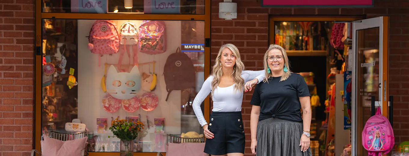 Två glada kvinnor står utanför en butik med fönsterskyltning med leksaker och accessoarer i rosa och vita färge