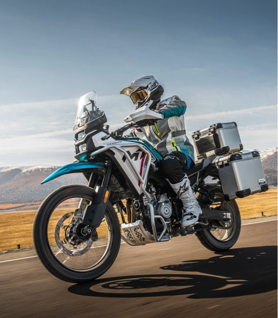 Motorcyklist med packväskor kör på landsväg med fjäll och öppet landskap i bakgrunden.