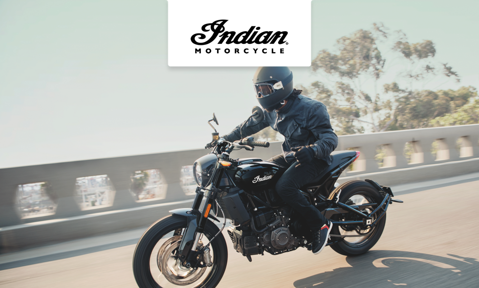 Förare på svart Indian-motorcykel kör längs solig väg med träd och bro i bakgrunden.