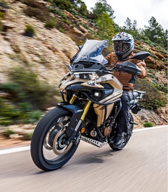 Motorcyklist kör en äventyrsmotorcykel på en slingrande bergsväg med klippor och grönska i bakgrunden.