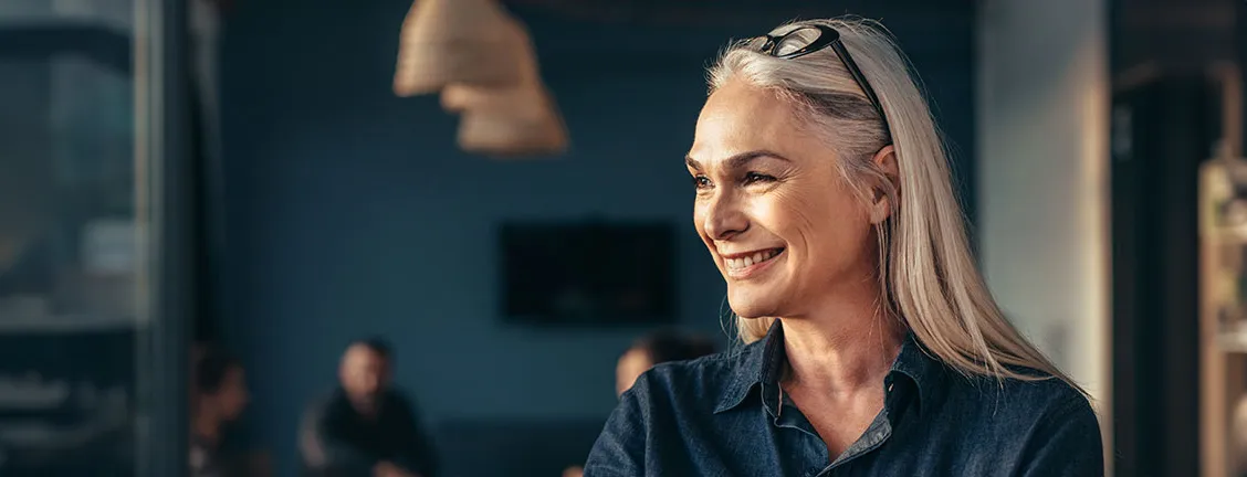 En leende äldre kvinna med grått hår och solglasögon uppdragna i håret ser åt sidan i en inomhusmiljö med svagt ljus och suddiga personer i bakgrunden