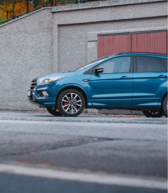 En blå SUV parkerad vid sidan av en gata med röd garagedörr och grå väggar i bakgrunden, tydliga röda och vita reflexer på hju